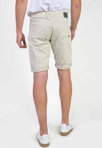 Shorts en denim beige avec des revers, dotés de poches arrière et d'un détail en patch noir. Portés avec des baskets blanches, affichant une texture lisse.
