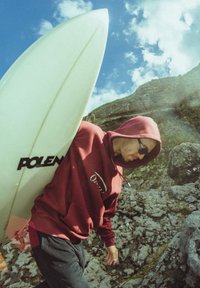 Planche de surf avec une finition en fibre de verre vert pâle, portée par une personne en sweat à capuche bordeaux et pantalon noir, marchant sur un terrain rocheux.