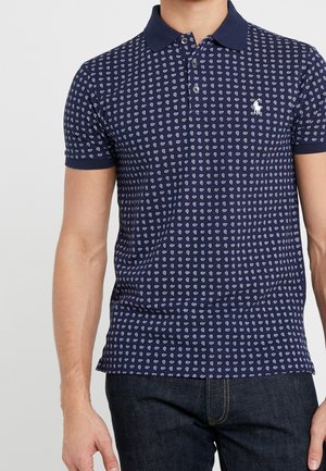 Man draagt donkerblauw poloshirt met klein wit geometrisch patroon en donkere spijkerbroek, staand tegen een effen lichte achtergrond.