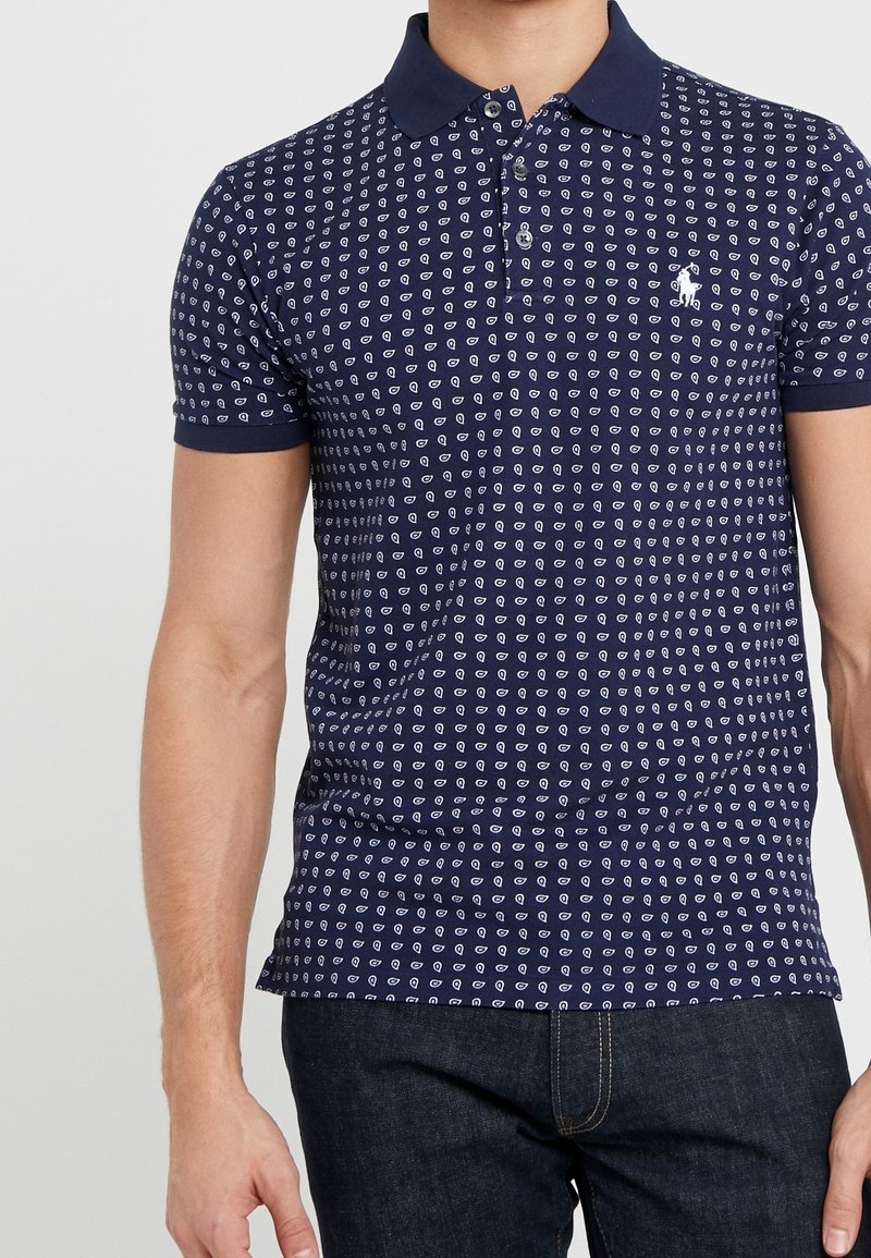 Homme portant un polo bleu marine avec un petit motif géométrique blanc et un jean foncé, debout devant un fond clair uni.