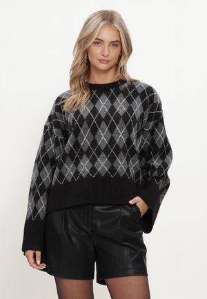 Frau mit blondem Haar, die einen schwarz-grauen Argyle-Pullover und schwarze Leder-Shorts trägt, steht vor einem schlichten weißen Hintergrund.