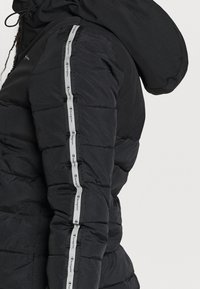 Svart puffjacka med kviltad design, vertikala Champion-logor på sidorna och en huva för extra isolering.