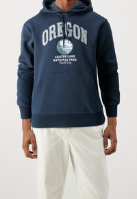 Tmavě modrá mikina s kapucí a přední kapsou ve stylu kengury, s grafikou Oregon a textem "NÁRODNÍ PARK KRÁTEROVÉHO JEZERA". Ribrový lem a manžety.