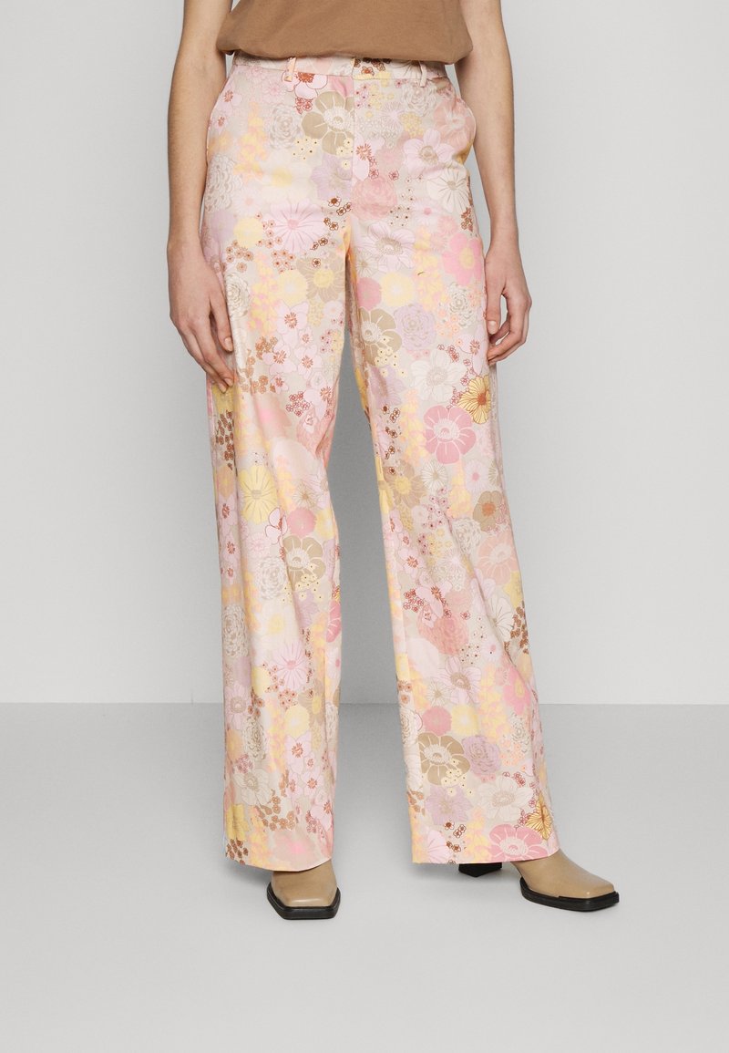 Femme portant un pantalon large taille haute à imprimé floral pastel et des bottes beiges, debout devant un fond uni gris clair.