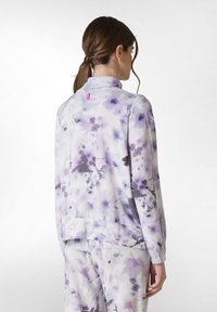 Lila blommönstrad jacka gjord av mjukt tyg med hög krage och rosa etikett detalj på baksidan, matchad med likadana byxor.