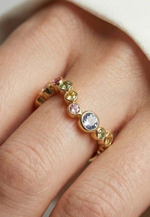 Guld ring med rundt farverigt ædelsten i forskellige størrelser, båret på en finger med lys hud.
