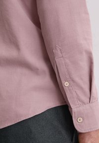 Jasnoróżowa koszula zapinana na guziki o gładkiej fakturze, z długimi rękawami, zaokrąglonym dołem i mankietami z dwoma guzikami, w połączeniu z ciemnoszarym materiałem poniżej.