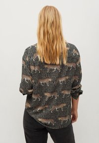 Schwarze Bluse mit Leopardenmuster und kleinen weißen Punkten, mit langen Ärmeln und lockerem Schnitt. Die Rückansicht zeigt fließenden Stoff und kontinuierliches Muster.