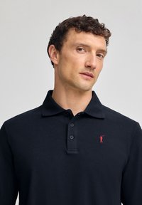 Polo shirt nero a maniche lunghe in un tessuto testurizzato con abbottonatura a tre bottoni e un piccolo logo ricamato rosso sul lato sinistro del petto.