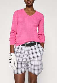 Mulher vestindo um suéter de malha canelada longo em rosa brilhante, calções à medida em xadrez branco e preto com um cinto preto e uma luva de golfe branca na mão esquerda.