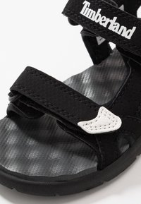 Czarna sandał z paskami na rzep, teksturowaną szarą wkładką i białym przeszyciem jako akcentem. Posiada gumową podeszwę i logo Timberland.