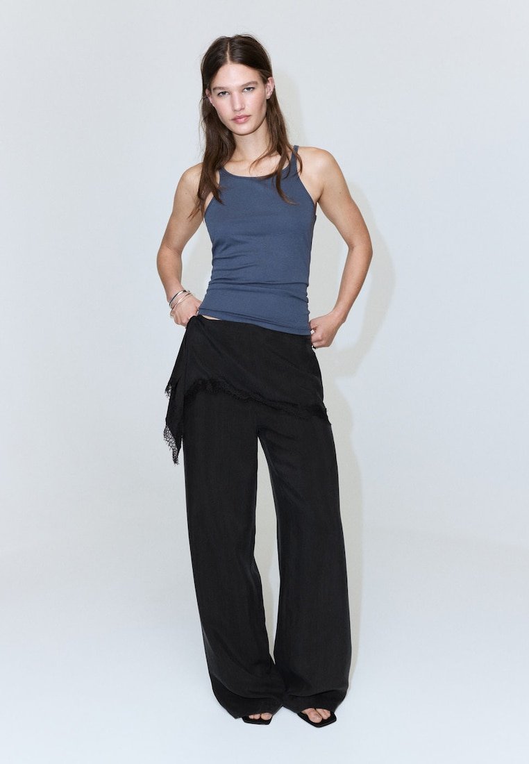 Jeune femme debout portant un haut sans manches bleu, un pantalon noir à jambes larges et une jupe portefeuille asymétrique noire.