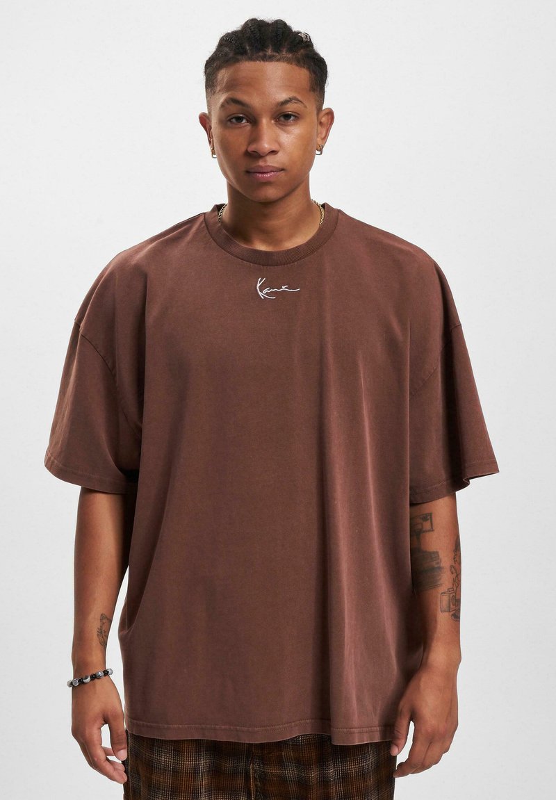 Brown oversized cotton t-shirt with a round neckline, featuring a white embroidered logo on the chest. Short sleeves and a relaxed fit.