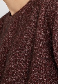 Pull en maille texturée de couleur bordeaux foncé, avec un col rond et un léger éclat agrémenté d'un motif moucheté.