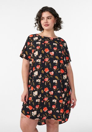 Mujer con cabello rizado que lleva un vestido negro de manga corta con flores rojas, blancas y beige sobre un fondo liso.