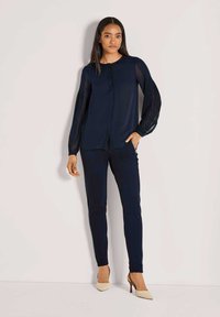 Marinebluse mit gefalteten, durchsichtigen Ärmeln, Knopfleiste und passenden marineblauen, figurbetonten Hosen. Das Model trägt beigefarbene High Heels. Minimalistisches Design.