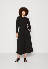 Abito lungo nero con maniche lunghe, chiusura con bottoni, vita cintrata e tasche, realizzato in tessuto leggero. Colletto arrotondato, design lungo fino alla caviglia.