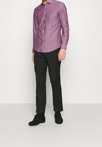 Muž v dlouhém rukávu fialové košili na knoflíky, černých kalhotách a černých společenských botách stojící proti jednoduchému bílému pozadí.