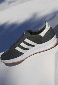 Zapatilla deportiva en blanco y negro con tres rayas blancas, suela de goma y talón con marca, mostrada sobre un fondo difuminado azul y blanco.