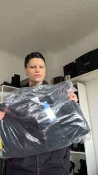 Peça de roupa preta segurada numa bolsa de plástico transparente, com um rótulo impresso. Textura mate, bordas lisas, sem padrões ou acessórios visíveis.