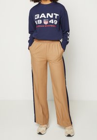 Námořnický mikina s dlouhým rukávem s velkým bílým a červeným potiskem "GANT 1949". Béžové široké kalhoty s námořnickými bočními pruhy. Ležérní sportovní oblečení.