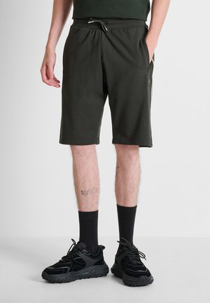 Uomo che indossa pantaloncini sportivi neri, calzini neri alti e scarpe da corsa nere, in piedi con le mani rilassate, una in tasca, su un pavimento grigio chiaro.