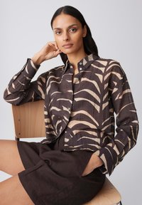 Woman seated on wooden chair wearing a dark brown and beige patterned shirt with dark brown shorts, looking at the camera with hand near face.