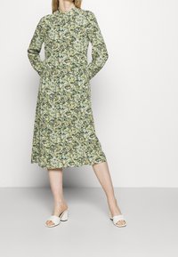 Blumenmusterkleid mit langen Ärmeln und hohem Ausschnitt, mit einem grünen und beigen Muster. Kombiniert mit weißen, mit Absatz ausgestatteten Mules, vor einem hellen Hintergrund stehend.
