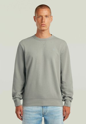 Uomo con capelli corti che indossa una felpa grigia chiara girocollo e jeans azzurri, in piedi davanti a uno sfondo beige uniforme.