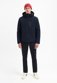 Marineblauwe geïsoleerde jas met capuchon, voorzijde met rits en gele accenten. Gecombineerd met donkere broeken en witte sneakers. Rood gebreide muts draagt men.