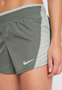 Pantalones cortos deportivos de Nike, con un cuerpo en verde oliva, paneles laterales de malla gris claro con pequeñas perforaciones y un logo bordado.