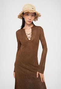 Abito marrone lavorato a maglia con maniche lunghe e scollo a V profondo con dettaglio incrociato. Indossato con un cappello di paglia naturale con frange.