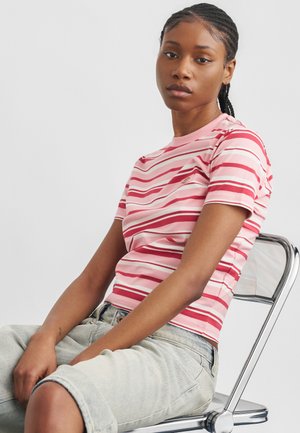 Νεαρή γυναίκα με πλεξούδες, φοράει ριγέ ροζ και κόκκινο μπλουζάκι και ανοιχτόχρωμα τζιν σορτς, κάθεται σε μεταλλική καρέκλα μπροστά σε λευκό φόντο.