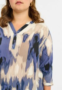 Blusa blu e beige con motivo, maniche a tre quarti, scollatura a V, patta dei bottoni e una texture del tessuto liscia.