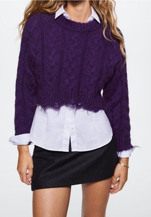 Femme portant un pull court en maille torsadée violet par-dessus une chemise blanche boutonnée et une mini-jupe noire ajustée, debout les bras détendus.