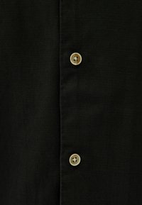 Tessuto nero testurizzato con due bottoni rotondi beige cuciti verticalmente sulla patta frontale di un indumento.