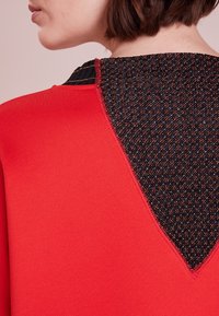 Suéter rojo con un respaldo de tejido negro texturizado y motas multicolores. El escote presenta un detalle de canalé en contraste.