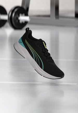 Μαύρο και τιρκουάζ παπούτσι τρεξίματος Puma με λευκή σόλα, αιωρούμενο πάνω από αντανακλαστικό πάτωμα με θολά γυμναστηριακά βάρη στο φόντο.