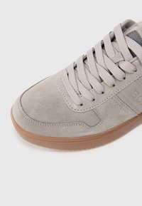 Zapatilla de ante gris claro con suela de goma beige, cordones planos y detalle de logo lateral. Textura suave y diseño de puntera redondeada.