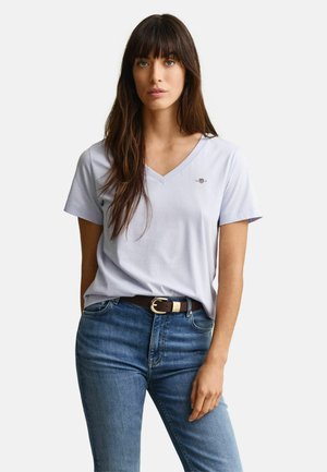 Vrouw met lang bruin haar, gekleed in een lichtblauw V-hals T-shirt dat in een blauwe spijkerbroek met een donkere riem is gestopt, tegen een effen achtergrond.