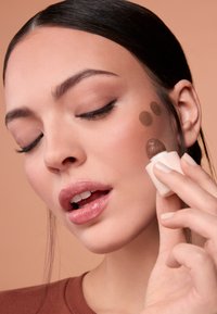 Aplicador de maquillaje sostenido cerca de la mejilla con tres círculos de producto marrón. La modelo tiene labios brillantes, maquillaje de ojos natural y piel suave.