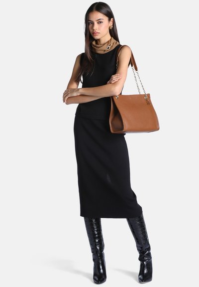 Femme portant une robe noire sans manches et des bottes noires montant jusqu'aux genoux, portant un grand sac à main en cuir marron avec des anses chaînes dorées sur l'épaule.