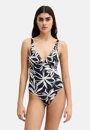 Mujer con traje de baño de una pieza negro con estampados de hojas tropicales blancas y tirantes finos, posando frente a un fondo claro y liso.