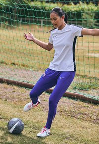 Balta Nike trumpa rankovė marškinėliai su juodais akcentais, purpurinės sportinės kelnės ir balti/raudoni futbolo batai, stovintys šalia juodo futbolo kamuolio.