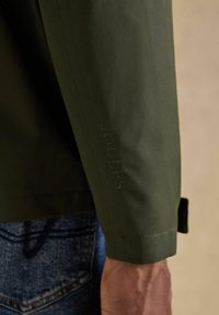 Nahaufnahme eines Arms einer Person, die eine olivgrüne Jacke mit dem auf dem Ärmel geprägten Schriftzug "Joules" trägt, kombiniert mit blauen Jeans.