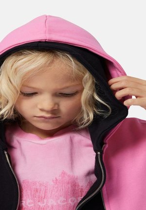 Felpa nera e rosa con cappuccio e zip frontale, morbida al tatto, con cappuccio regolabile, indossata sopra una t-shirt grafica rosa.