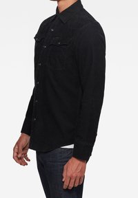 Czarna koszula z materiału corduroy z kołnierzykiem zapinanym na guziki, dwoma kieszeniami na piersi i długimi rękawami. Posiada widoczne szwy i metalowe guziki.