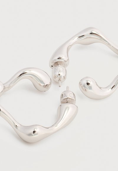 Ασημένια μεταλλικά σκουλαρίκια κρίκοι με κυματιστή μορφή και κούμπωμα πίσω με καρφί, τοποθετημένα σε λευκό φόντο.