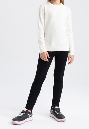 Dziecko ubrane w biały sweter, czarne dopasowane spodnie i czarne trampki z białymi i różowymi podeszwami stoi na jednolitym tle.