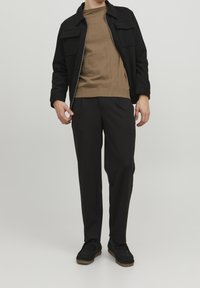 Giacca nera con zip e due tasche sul petto, maglione in maglia marrone e pantaloni neri. Le calzature sono nere con una suola texturizzata color nocciola.